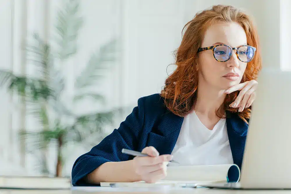 young woman with red hair and glasses working at a computer