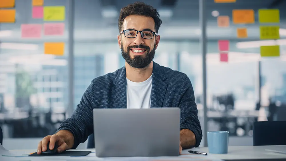 smiling young man with a beard and glasses looking up at the camera from his laptop