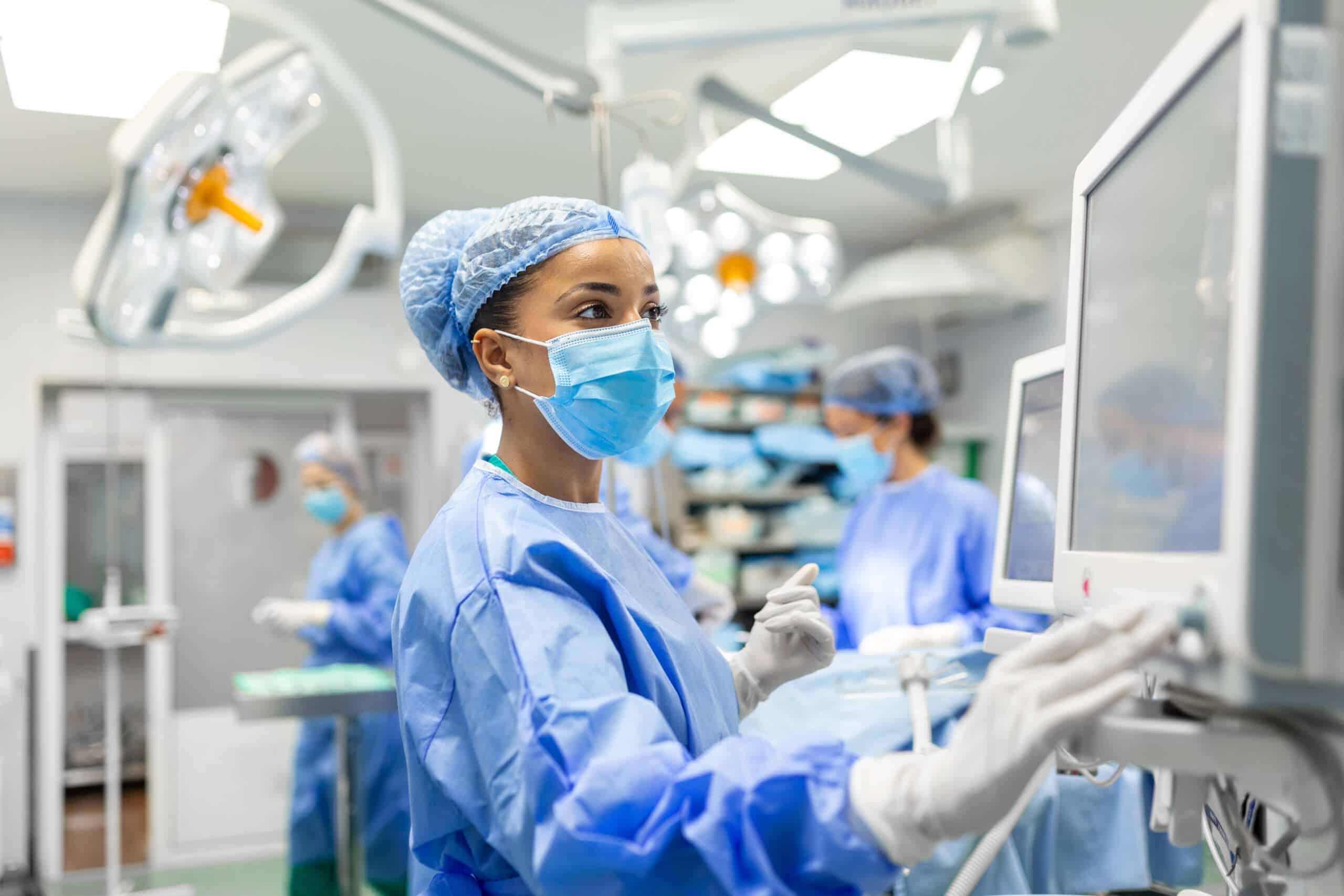 Anesthetist Working In Operating Theatre Wearing Protecive Gear checking monitors while sedating patient before surgical procedure in hospital